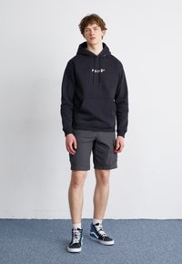 Sudadera negra con logo blanco en el pecho, combinada con pantalones cortos de carga grises. El modelo lleva zapatillas altas negras y azules. Fondo simple.