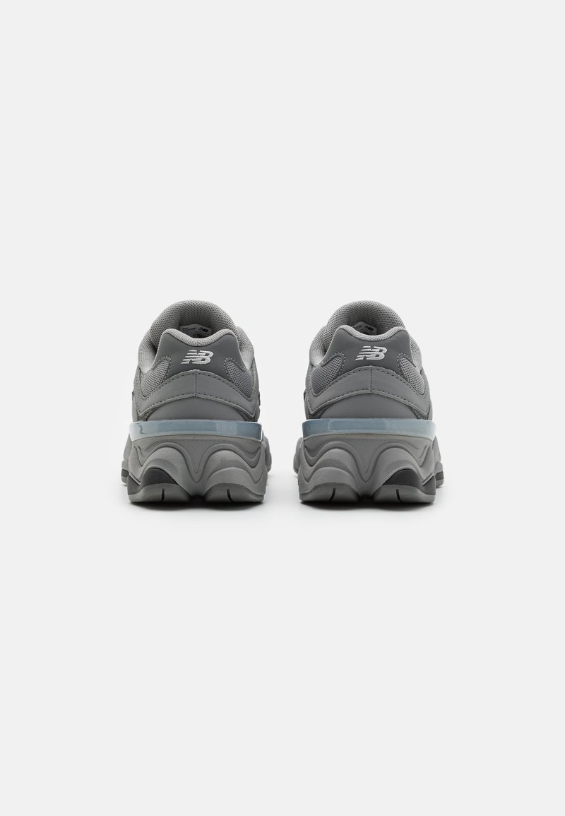Ein Paar graue New Balance Sneaker von hinten auf einem weißen Hintergrund gezeigt, die dicke Sohle und Netzdetails hervorhebend.