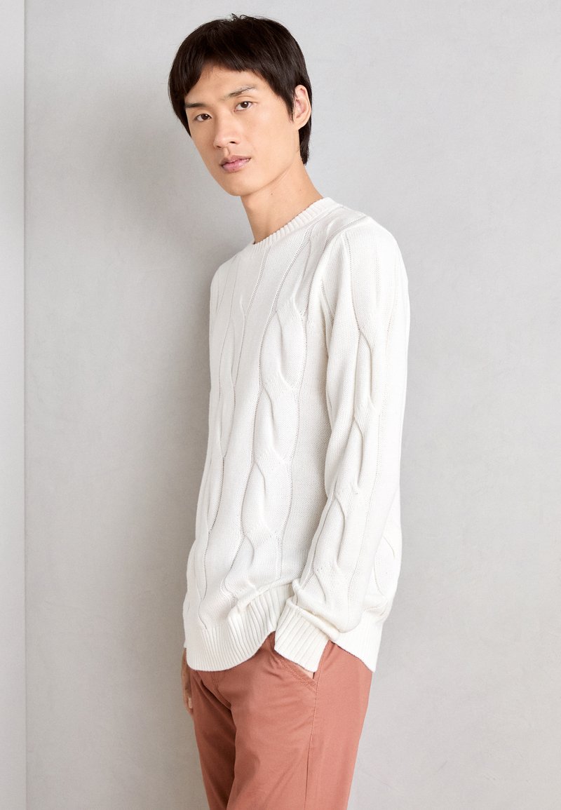 Valkoinen kaapelikudottu neulepaita, jossa on pyöreä kaula-aukko, tekstuurimallinen pinta ja ribbireunat, yhdistettynä vaaleanpunaisiin housuihin.