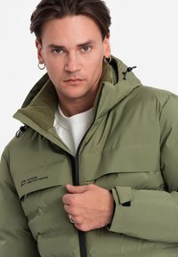 Olivově zelená péřová bunda se zapínáním na zip, nastavitelnou kapucí a fleecovou podšívkou. Obsahuje logo značky a detail na manžetách.