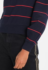 Suéter navy con rayas horizontales rojas, puños y dobladillo acanalados, textura suave. Combinado con pantalones negros con detalles de franjas laterales.