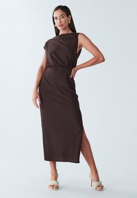 Robe midi marron côtelée sans manches avec une fente latérale et une taille froncée. Associée à des sandales à talons hauts tressées de couleur claire.