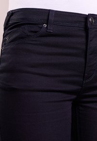 Mörka denimjeans med en slät textur, som har en figurnära passform, kontrasterande sömmar och en framficka med hårdvarudetaljer.