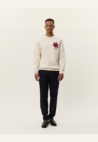 Sudadera crema con diseño de flor roja en el lado izquierdo del pecho, acompañada de pantalones azul oscuro y zapatos negros. Textura suave, escote redondo.