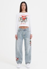 Hvid, langærmet crop top med grafisk print; lyseblå, løstsiddende jeans med broderede designs i rød og grøn; hvide sneakers.