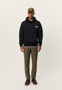 Μαύρο φούτερ με κουκούλα και λευκό λογότυπο "LES DEUX", τσέπη καγκουρό· παντελόνι σε ελιά, εφαρμοστό· καφέ παπούτσια. Καπέλο με λεπτομέρεια "II" σε ελιά.