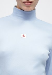 Φωτεινό γαλάζιο ριπ ζιβάγκο τοπ με ένα εμφανές τετράγωνο λογότυπο σε κόκκινο και λευκό, με το κείμενο «Calvin Klein» κοντά στη γραμμή του λαιμού.