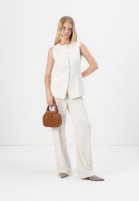 Gilet beige senza maniche con chiusura a bottoni, abbinato a pantaloni larghi coordinati. Tiene in mano una piccola borsa marrone con texture e indossa scarpe beige a punta.