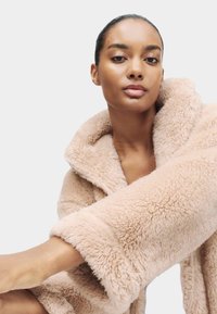 Donna con la pelle liscia e i capelli raccolti, che indossa un morbido cappotto beige e peloso su uno sfondo leggero e neutro.
