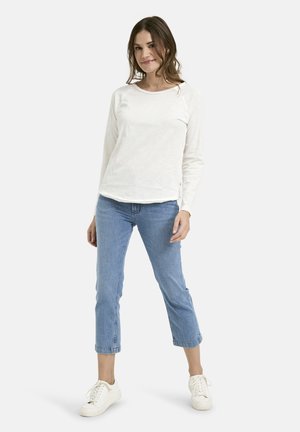 Frau steht und trägt ein langärmliges weißes Hemd, hellblaue Jeans in Cropped-Länge und weiße Turnschuhe vor einem schlichten weißen Hintergrund.