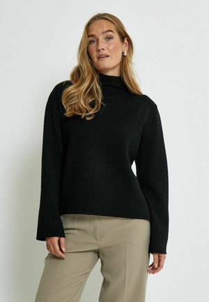 Zwarte coltrui met lange, uitlopende mouwen en een relaxed fit, gemaakt van een gladde, zachte stof. Gestyled met beige broek.
