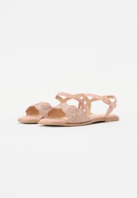 Rosa platta sandaler med blommig spetstrykt framremmar och anklar med spännen på en enfärgad vit bakgrund.