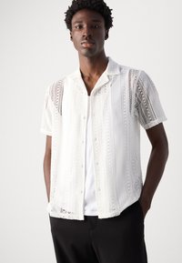 Cămașă albă cu mânecă scurtă, cu un design din dantelă cu modele intricate, guler și închidere cu butoane, suprapusă peste un tricou alb simplu.