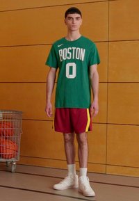 Grøn Nike T-shirt med "BOSTON" og nummer "0", røde shorts med gule striber, hvide sneakers, stående i et træningsmiljø.