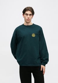 Dark green long-sleeve sweatshirt made of cotton, featuring a small yellow graphic logo on the left chest area. Relaxed fit with ribbed cuffs.
