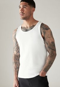 Weißes Baumwoll-Top mit Rundhalsausschnitt, verziert mit filigranen Tattoo-Designs am Oberarm, vor einem neutralen Hintergrund.