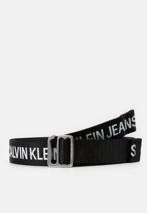 Sort stofbælte med sølvspænde, hvidt Calvin Klein Jeans-logo trykt langs hele længden, vist sammenrullet på en ensfarvet baggrund.