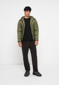 Jachetă pufoasă verde măsliniu, cu glugă, fermoar frontal și manșete elastice, purtată peste un tricou negru și pantaloni închiși la culoare, asortată cu încălțăminte neagră.