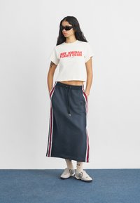 Κομμένη κρεμ μπλούζα με κόκκινο κείμενο "AIR JORDAN FIGHT CLUB", σε συνδυασμό με σκούρα μπλε φούστα με κόκκινες και λευκές πλευρικές ρίγες. Φορεμένη με γυαλιά ηλίου και αθλητικά παπούτσια.