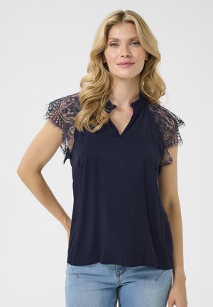 Haut bleu marine avec des manches en dentelle, décolleté en V et coupe détendue. Tissu doux avec un détail de col volanté. Associé à un jean bleu clair.