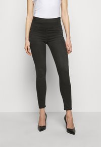 Jeans pitillo negros con una cintura alta, con bolsillos decorativos con cremallera y una textura de mezclilla suave y elástica. Combinados con tacones negros.