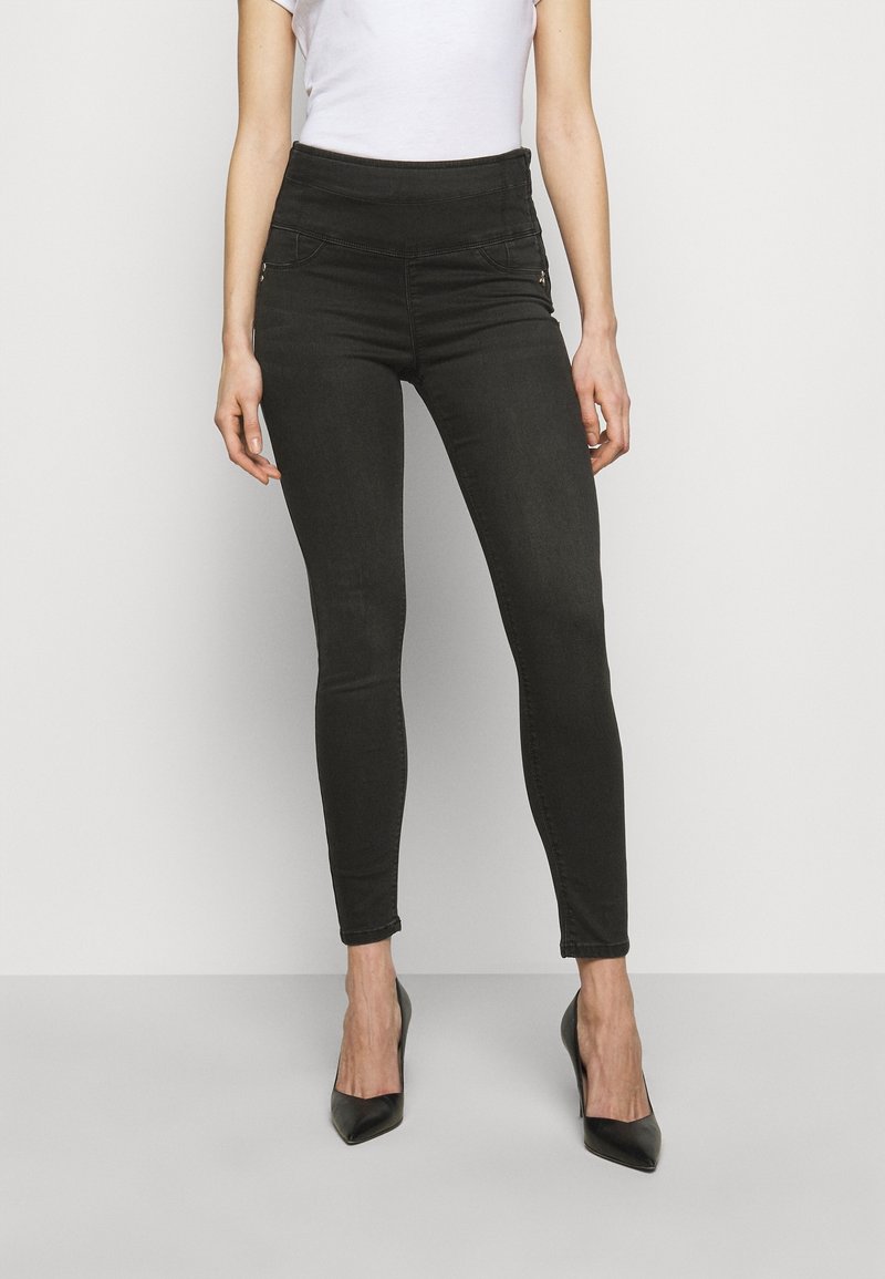 Jeans pitillo negros con una cintura alta, con bolsillos decorativos con cremallera y una textura de mezclilla suave y elástica. Combinados con tacones negros.