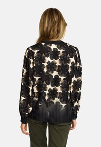 Kvinne med langt hår iført en svart og beige blomstrete langermet bluse og olivengrønne bukser, sett bakfra mot en ensfarget bakgrunn.