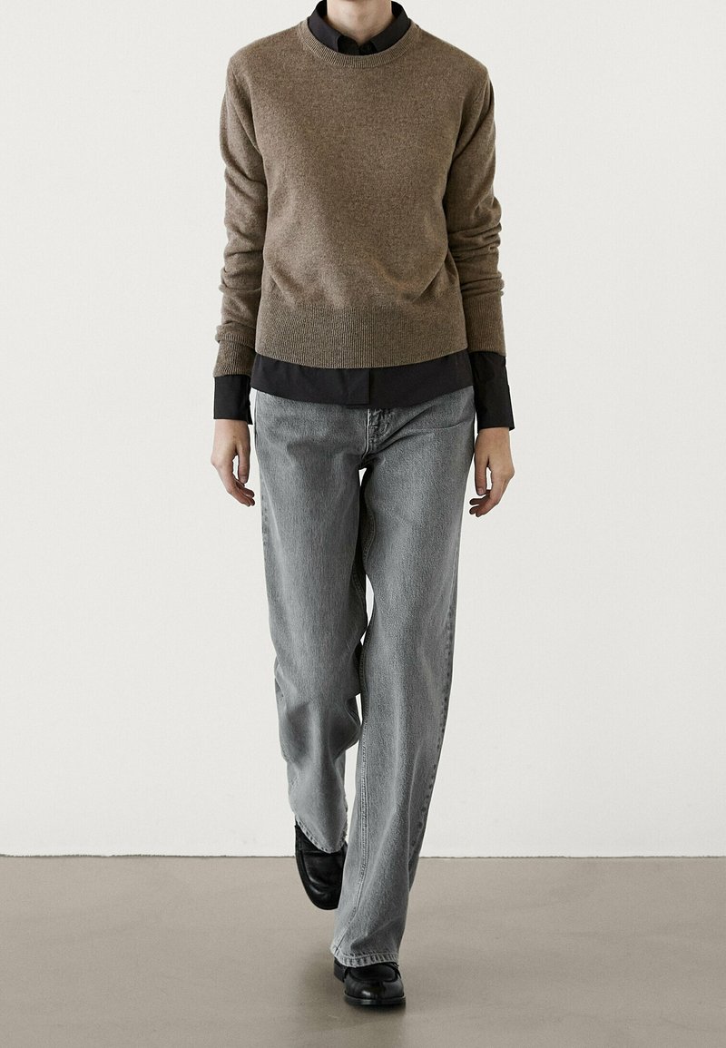 Pull en maille marron par-dessus une chemise noire à col, associé à un jean large gris clair et des chaussures noires, sur un fond minimaliste.
