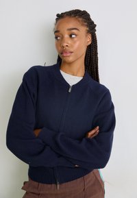 Jūrinės vilnos zip-up megztinis su raukiniais ant rankovių ir apykaklės, turintis apvalią kaklo iškirpę ir raglan tipo rankoves. Lygi tekstūra ir prigludusi forma.
