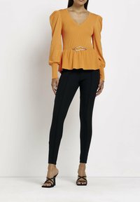 Top peplum a coste arancione con scollo a V, maniche a sbuffo e un accento in metallo argentato in vita, abbinato a pantaloni neri aderenti e tacchi neri.