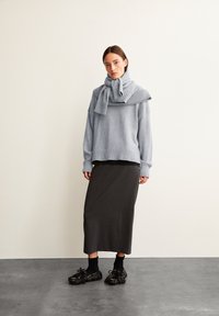 Lichtgrijze gebreide trui met sjaaldetail, gecombineerd met een donkergrijze midi-jurk en zwarte schoenen met franjes. Soepele texturen.