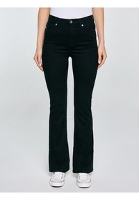 Sieviete, kas valkā melnas bootcut džinsas un baltus sporta apavus, stāv ar rokām brīvi nokarenām gar sāniem pret vienkāršu fonu.