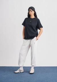 Juoda marškinėliai su mažu baltu „Nike“ logotipu, pilkos sportinės kelnės ir smėlio spalvos sportbačiai su dekoratyviniu numeriu šone; kepurė dėvima atgal. Esk