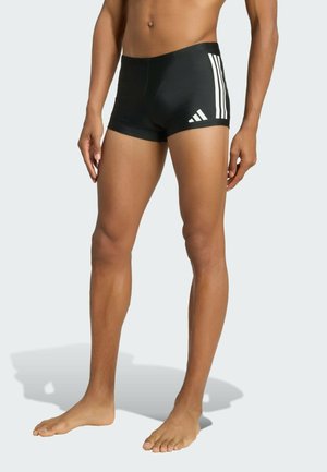 Mężczyzna w czarnych kąpielówkach z białym logo Adidas i trzema paskami na jasnym tle, stojący boso.