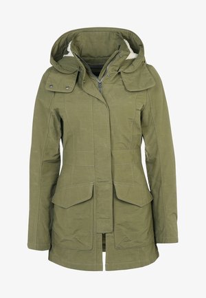 Veste imperméable vert olive avec capuche, associée à une jupe matelassée noire et des collants bordeaux ; des chaussures de sport noires complètent la tenue.