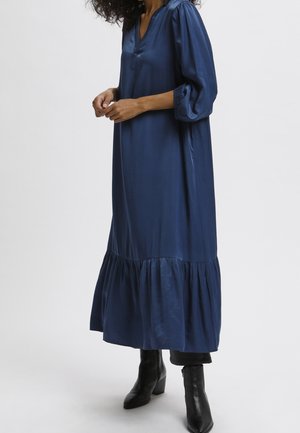 Frau trägt ein lockeres, langes dunkelblaues Kleid mit aufgeplusterten dreiviertel Ärmeln und schwarzen Stiefeletten mit Absatz.
