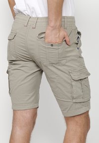 Bărbat îmbrăcat în pantaloni scurți bej cargo, cu mai multe buzunare, cu mâna bagată în buzunarul din spate, arătat de la talie până la genunchi pe un fundal simplu.