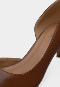 Καφέ αντλία με ψηλό τακούνι και λείο δερμάτινο εξωτερικό, στρογγυλεμένη μύτη και διακοσμητική ραφή κατά μήκος της άκρης. Ελαφρώς κυρτή σιλουέτα.