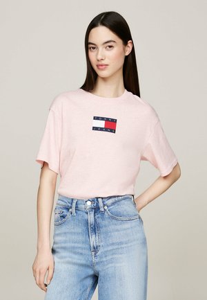 Camiseta de algodón rosa con mangas cortas y cuello redondo, que presenta un estampado rectangular de logotipo en azul marino y rojo en el frente. Jeans de mezclilla azul desgastados.