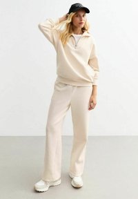 Tuta di pile beige con zip a metà e pantaloni con elastico in vita. Indossato con un cappello nero e scarpe sportive bianche.