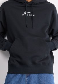 Svart Nike Air Max hoodie med en ficka fram, dragsko på huven och en vit logotyp på bröstet. Mjukt bomullsmaterial med ribbade muddkanter.