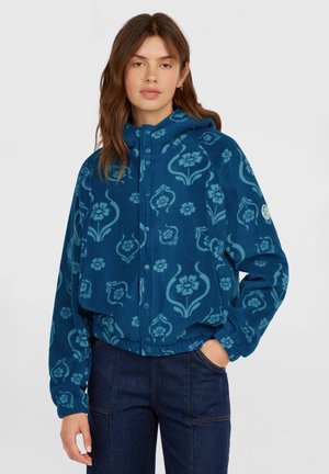 Jonge vrouw draagt een blauw fleecejack met patroon en capuchon en donkerblauwe broek, staand met één hand in de zak tegen een effen achtergrond.