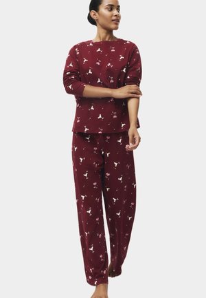 Una mujer luce un conjunto de pijama burdeos de manga larga, estampado con pequeñas copas de cóctel, sobre un fondo liso y claro.