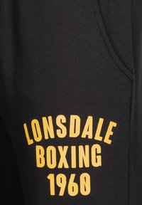 Melnas sporta bikses, kas izgatavotas no kokvilnas, ar mīkstu tekstūru. Uz tām ir dzeltena druka ar uzrakstu "LONSDALE BOXING 1960" treknos, arkas veida burtiem.