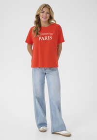T-shirt en coton rouge avec "MIDNIGHT IN PARIS" imprimé en blanc. Assorti avec un jean large bleu clair et des baskets beiges.