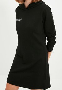 Svart hoodieklänning i smidigt tyg, med en kenguruficka och långa ärmar. Framsidan har en liten textdetalj i vitt.