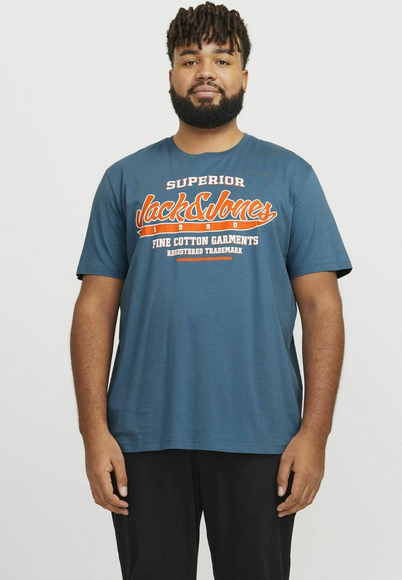 Camiseta de algodón azul con mangas cortas, que presenta un logo impreso en naranja y blanco en un diseño audaz. Cuello redondo, corte relajado, sin patrones ni detalles.