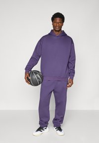 Paarse hoodie en bijpassende joggingbroek van zacht materiaal, met een comfortabele pasvorm. Het model houdt een basketbal vast en draagt zwart-witte sneakers.