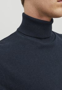 Pulover moale cu guler tip turtleneck în nuanță marină, fabricat dintr-un material tricotat, cu un guler cu nervuri și o textură netedă. Prim-plan al părții superioare a corpului și zonei gâtului.
