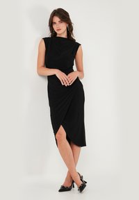 Čierne priliehavé šaty s drapovaným výstrihom a asymetrickým lemom, s bočným rozparkom. Modelka má na sebe čierne lodičky so špicatou špičkou.